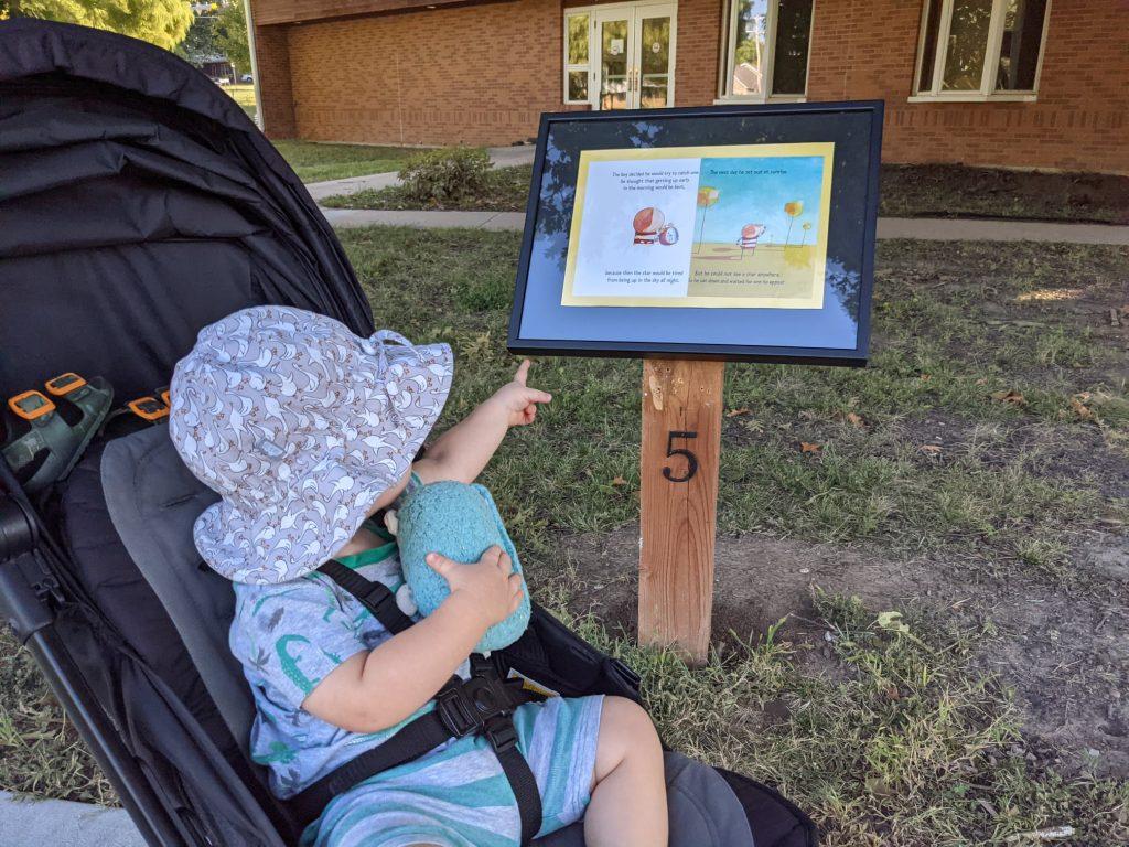 Boy in stroller points at StoryWalk board, which has two pages of a picture book on an outdoor stand.