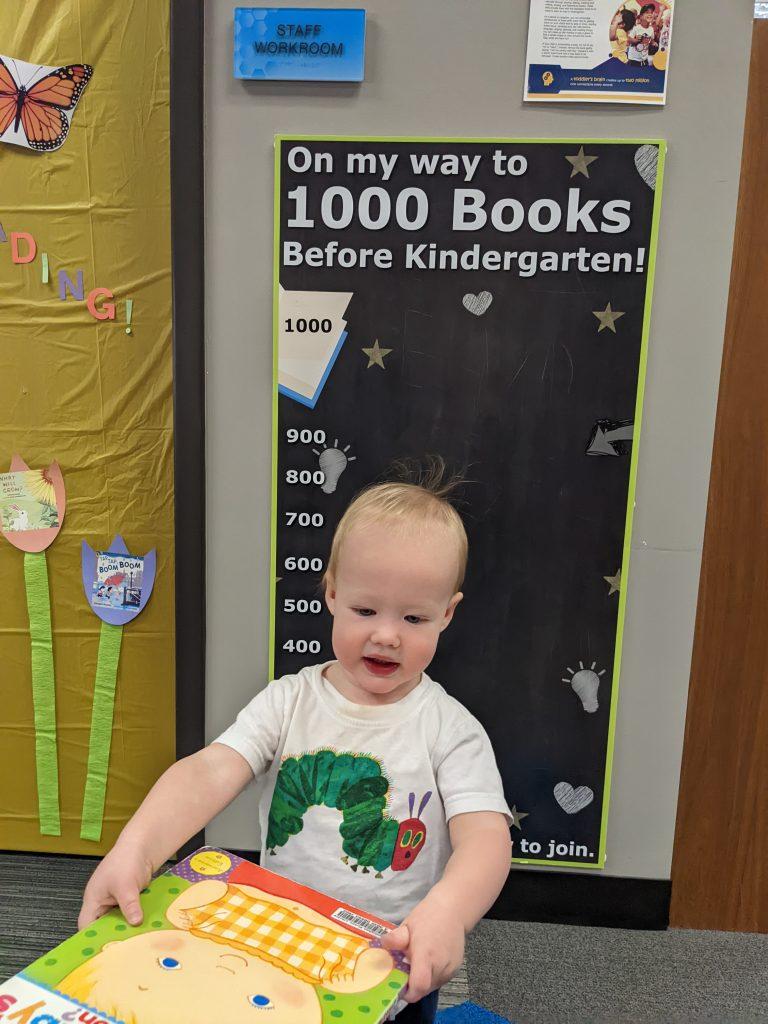 A toddler boy holds up a book in front of a large wall sign that says "On my way to 1000 Books Before Kindergarten!"