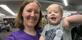 mom and toddler son wearing K-State shirts smile with Open House displays in the background