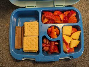 Segmented lunchbox containing cheese crackers, a Kit-Kat, strawberries, apples, bell peppers, and small meat snacks