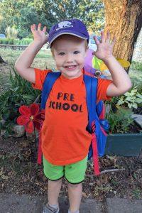 three-year-old boy taking a first day of school picture, wearing a t-shirt that says "preschool"