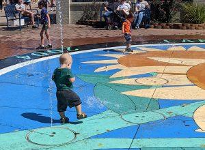 A toddler boy gets splashed in an outdoor fountain that spurts out of the ground